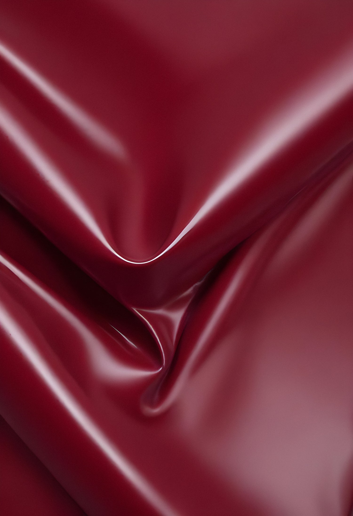 A close-up of deep red glossy leather fabric with soft, flowing folds illuminated by gentle light