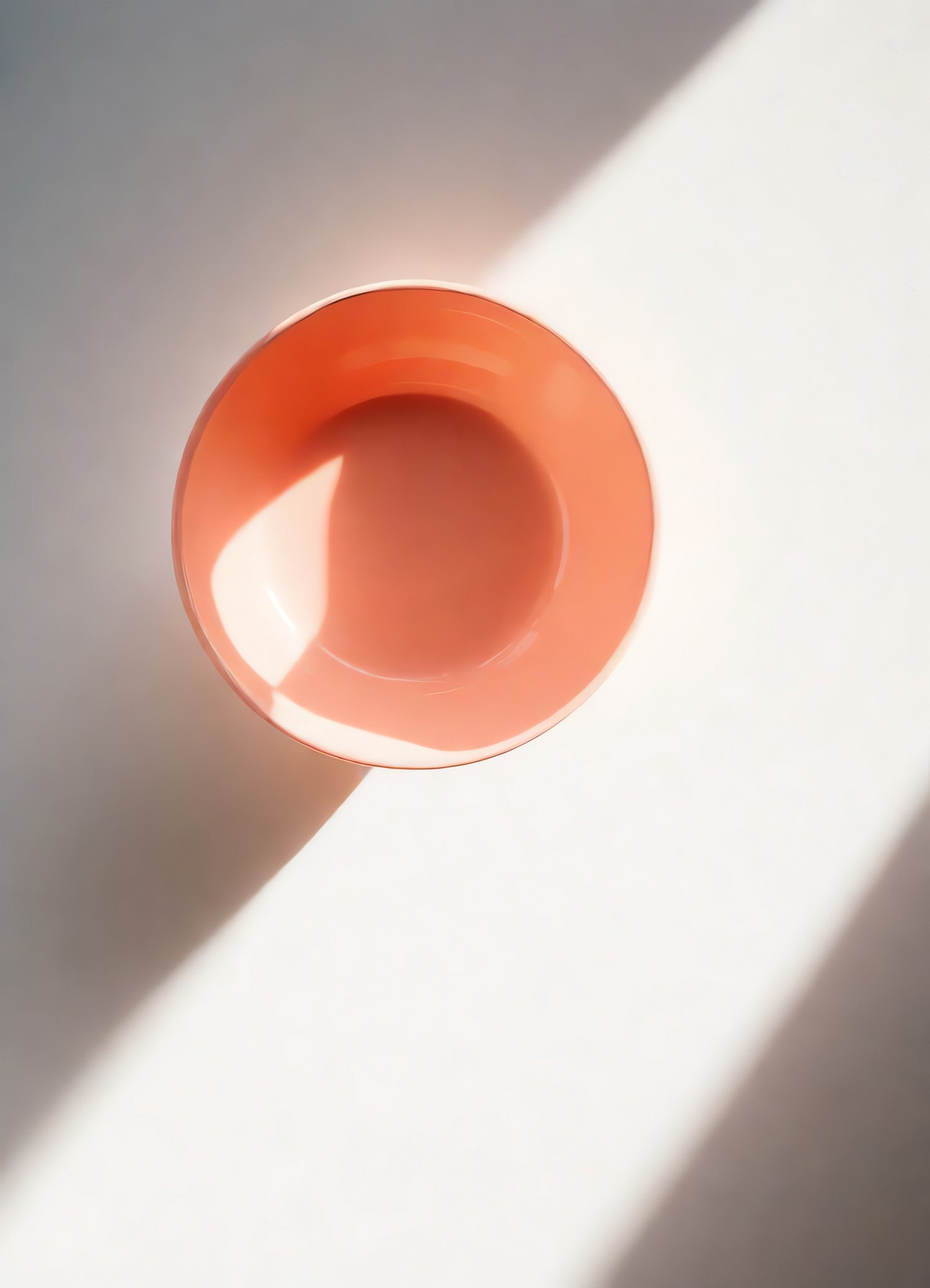 A simple orange bowl sits on a white surface illuminated by soft afternoon sunlight in a serene indoor setting