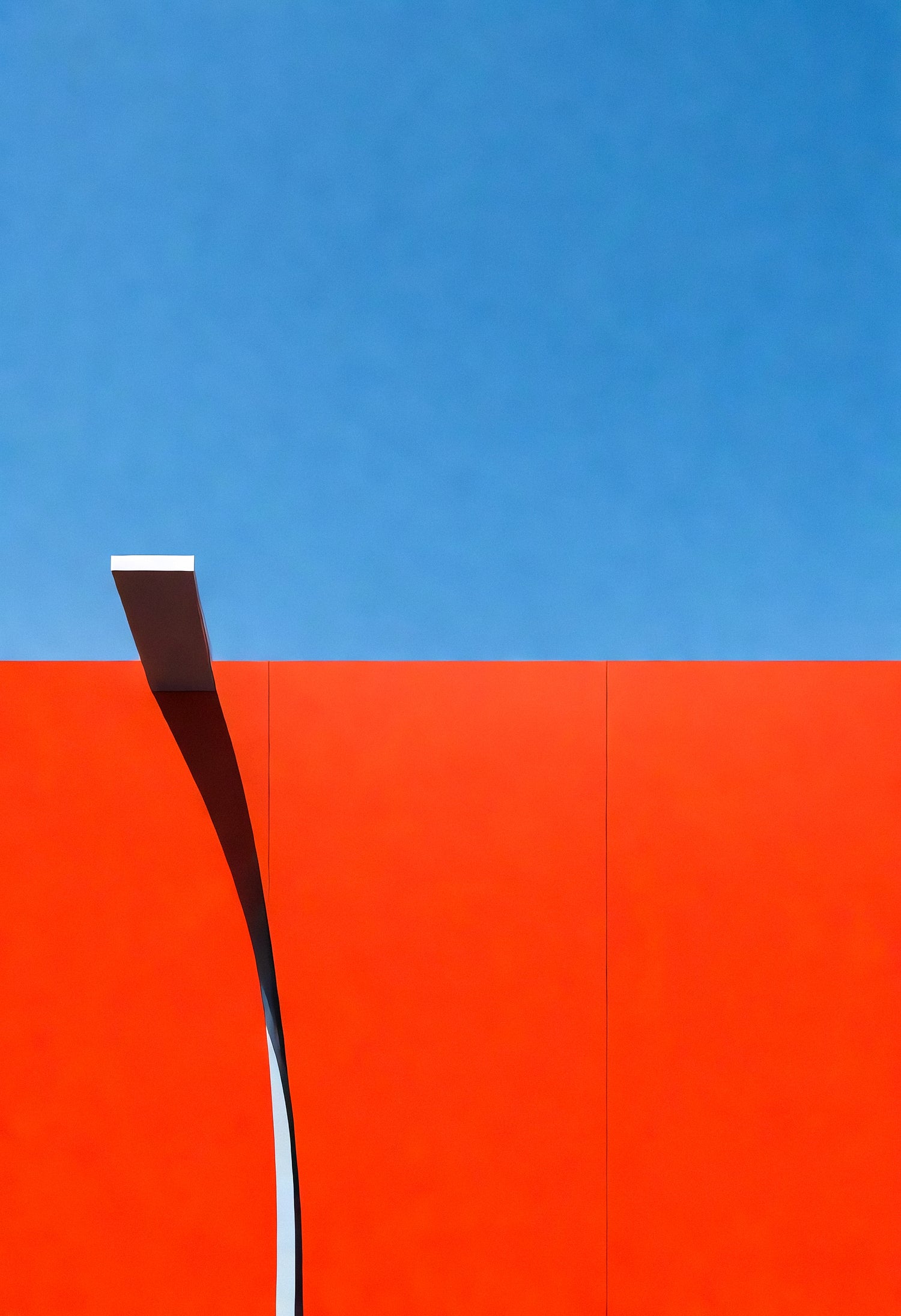 Modern architectural design featuring a curved light pole against a bright red wall and clear blue sky during the day