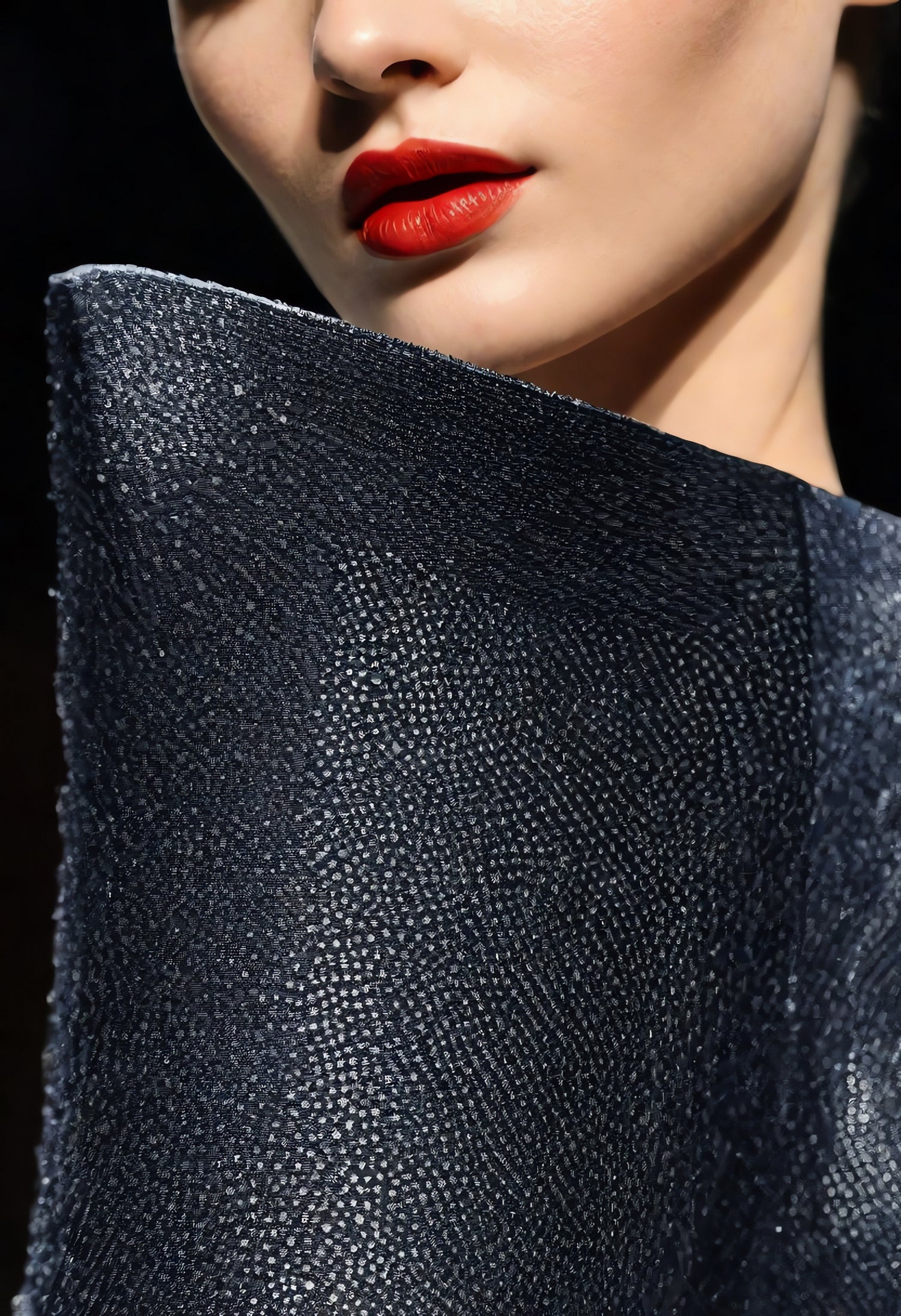 A close-up of a model showcasing a textured dark garment with bold red lipstick at a fashion event during evening hours